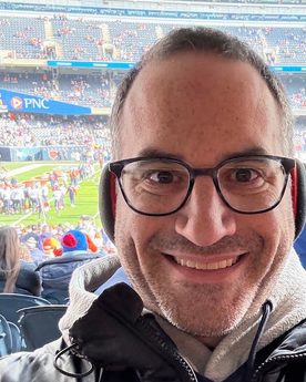 Zach Stern smiling at the camera, in glasses and a black winter jacket, in the stands at a football game with the field and the crowd behind him.
